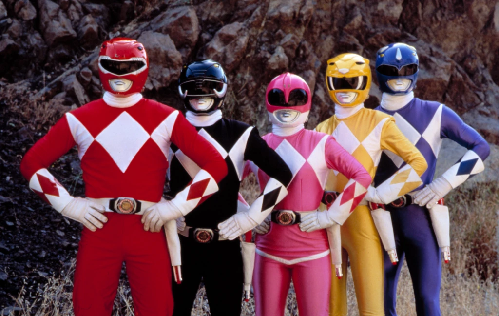 Five Power Rangers in red, black, pink, yellow, and blue costumes standing side by side with hands on hips outdoors against a rocky background.